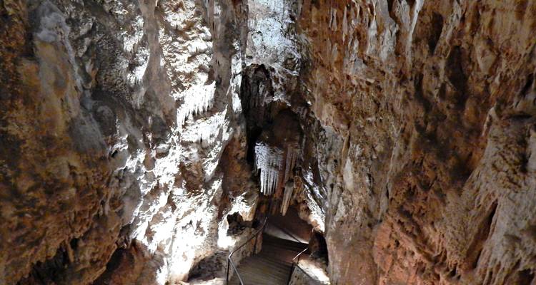Passage de grotte calcaire illuminé avec stalactites et passerelle en bois descendant dans l'obscurité.