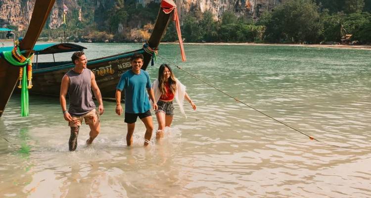 Three young adults wade through shallow turquoise water beside a decorated long-tail boat with dramatic limestone cliffs in the background.