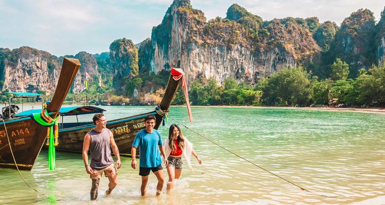 Trois voyageurs pataugent depuis des bateaux traditionnels à longue queue dans les eaux émeraude de la plage de Railay avec ses falaises de calcaire imposantes.