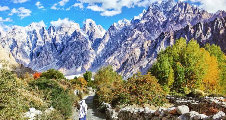 Persona caminando por un sendero rodeado de follaje colorido y montañas majestuosas.