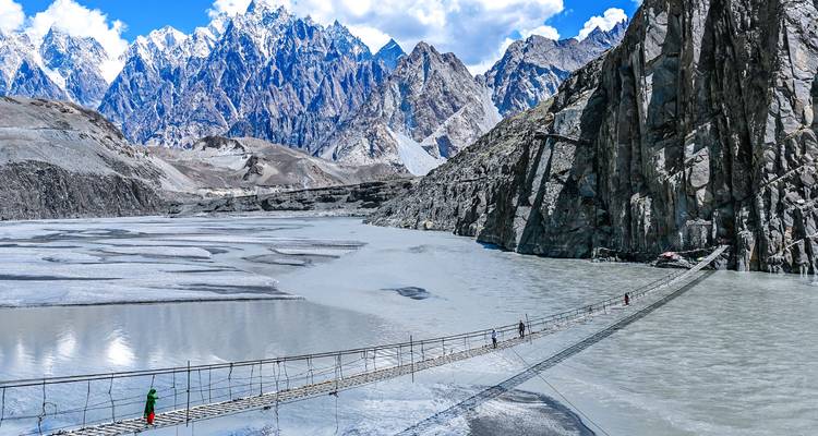 People crossing a suspension bridge over a river with mountains in the background.