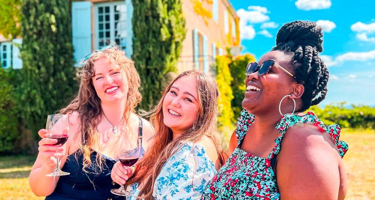 Trois femmes rient ensemble en tenant des verres de vin rouge près d'un château de campagne ensoleillé et des cyprès.