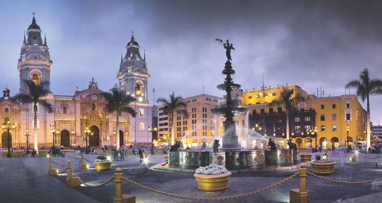 Avondaanzicht van Lima's Plaza Mayor met de verlichte kathedraal torens, koloniale gebouwen en centrale fontein onder een dramatische hemel.