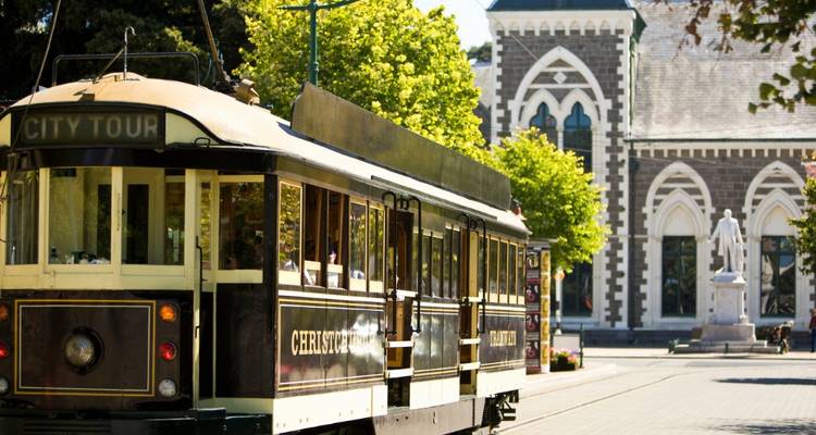 Vintage-Straßenbahn-Stadtführung durch Christchurch, die an einem gotischen Steingebäude auf einer baumbestandenen Straße vorbeifährt.