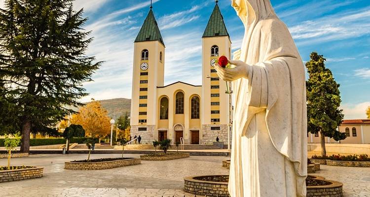 Statue holding a red rose in front of a church with two towers.