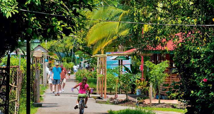 Ruelle de village tropical avec un enfant à vélo et des visiteurs marchant devant des maisons en bois colorées et des palmiers.