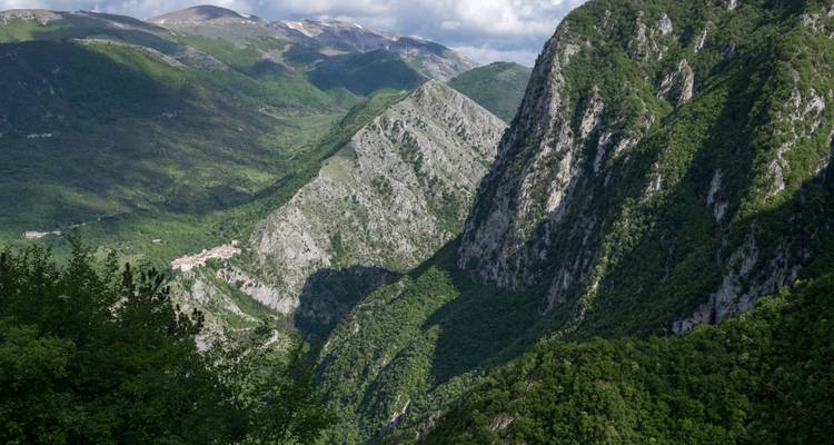 Deep green gorge with towering limestone cliffs and distant mountain peaks under a dramatic sky.