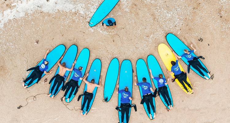 Een luchtfoto van surfleerlingen die liggen op felblauwe planken gerangschikt in een halve cirkel op een zandig strand.