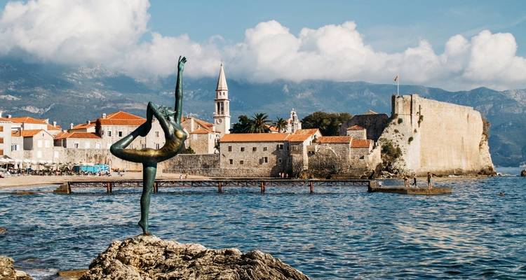 Statue einer Tänzerin auf den Felsen mit einer historischen Küstenstadt im Hintergrund.