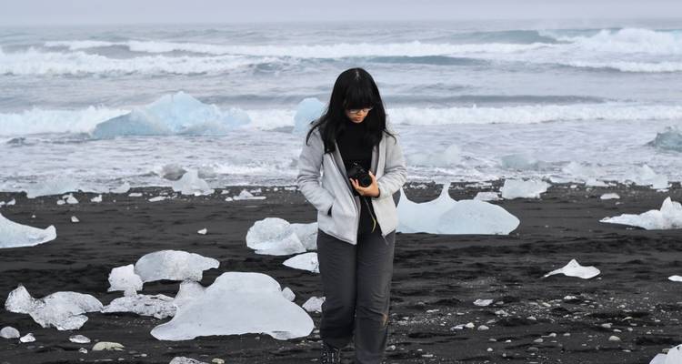 Young woman walks along black volcanic sand strewn with glacial ice chunks near the sea.