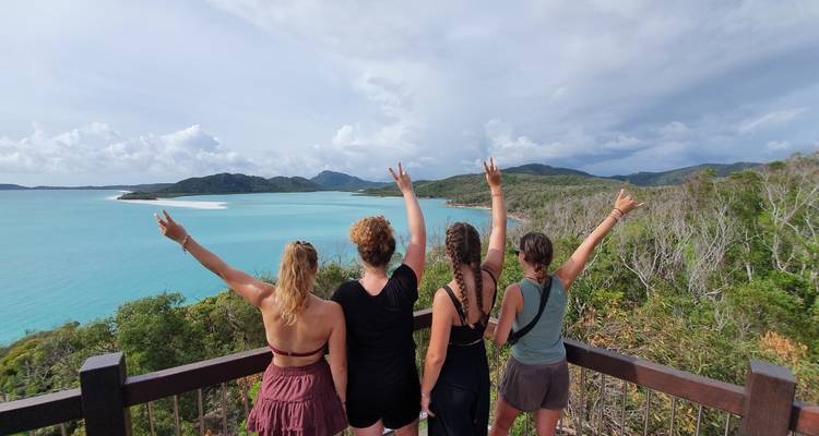 Four women facing a scenic ocean view with their arms raised.