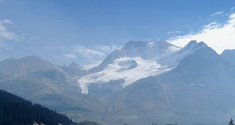 Entfernte, gletscherbedeckte Gipfel erheben sich über ein dunstig verschleiertes Tal unter einem blassen blauen Himmel.