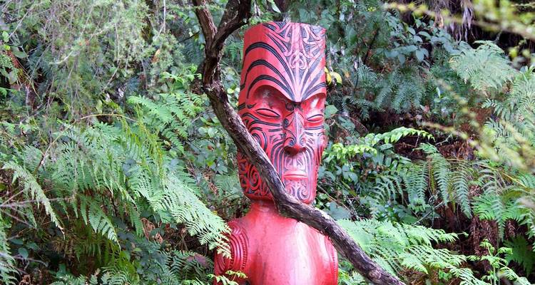 Eine traditionelle rote Maori-Schnitzerei steht inmitten üppiger grüner Farne und Waldvegetation.