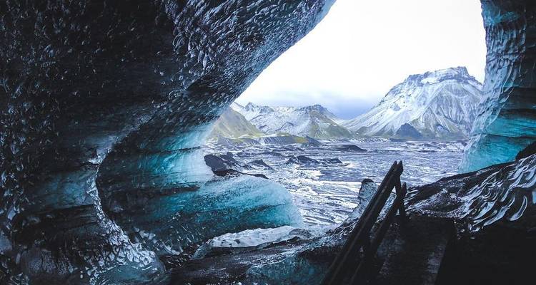 Ouverture de grotte de glace aux teintes bleues encadrant des montagnes escarpées enneigées et une vallée glaciaire.