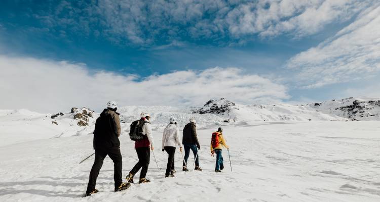Cinq randonneurs en raquettes traversent un vaste glacier blanc sous un ciel bleu éclatant et des nuages dispersés.
