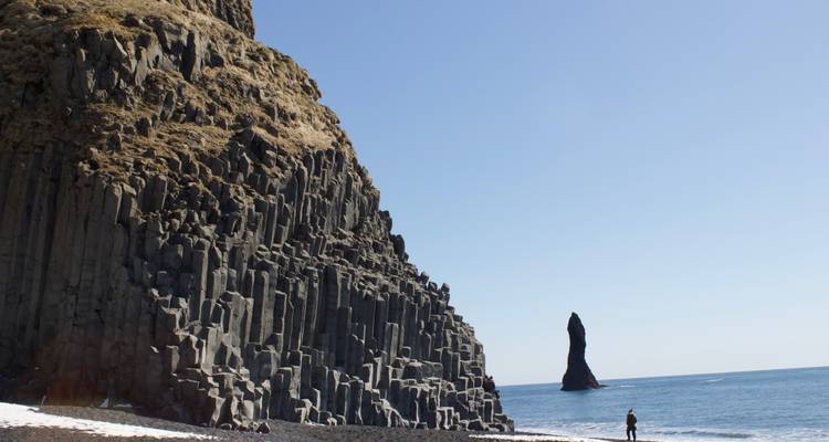 Un visiteur solitaire se tient sur une plage de sable noir à côté de colonnes de basalte imposantes et d'une formation rocheuse en mer au large.