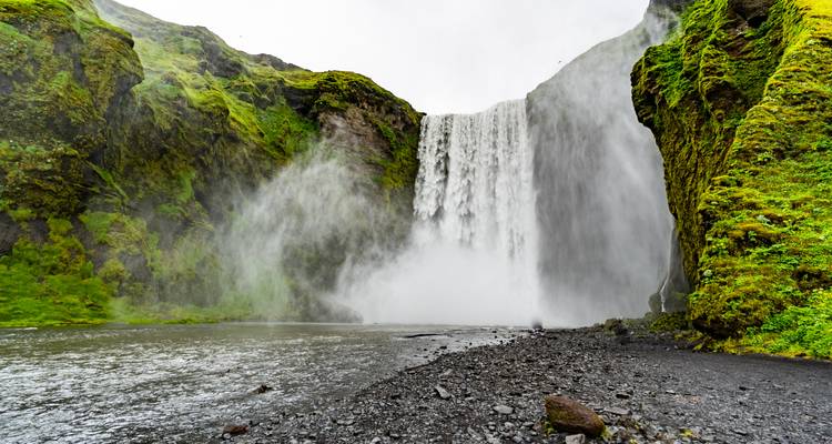 La puissante cascade de Skogafoss se jette sur des falaises vertes et mousseuses dans un bassin brumeux en contrebas.