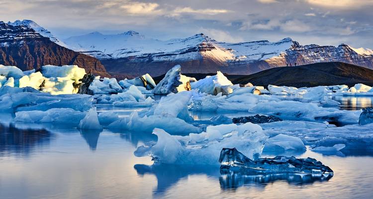 Des icebergs illuminés par le soleil flottent dans un lagon aux eaux cristallines avec des montagnes enneigées en arrière-plan.