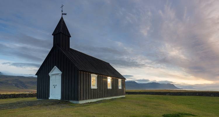 Une petite église en bois noir aux fenêtres illuminées se dresse sur un paysage herbeux dégagé, avec un arrière-plan montagneux islandais sous un ciel crépusculaire aux couleurs vives.