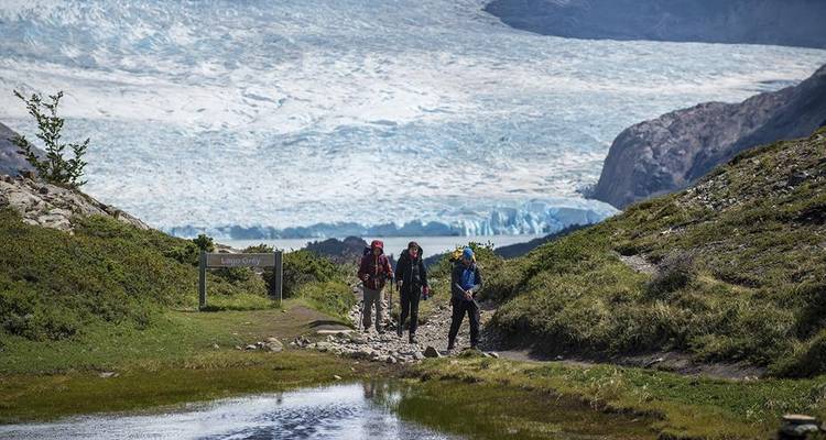 Drie wandelaars lopen over een rotsachtig pad naast een spiegelglad meer met een enorme blauwe gletsjer en ruige bergen die hoog boven hen uitsteken.