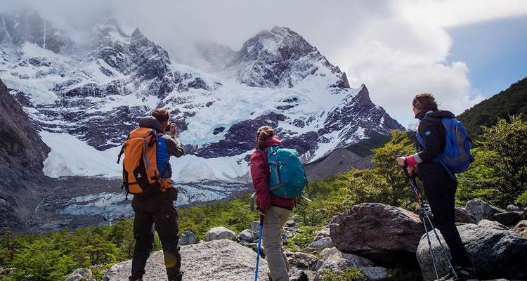 Drie trekkingwandelaars maken even pauze om de indrukwekkende ijzige pieken en hangende gletsjers boven een rotsachtige vallei te bewonderen.