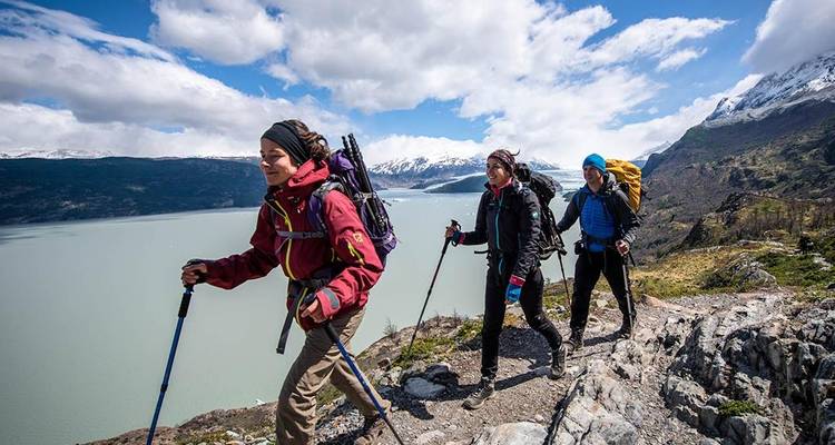 Wandelaars met trekkingstokken stappen flink voort langs een kliffrand die uitkijkt op een enorm grijs meer en sneeuwbedekte bergtoppen.