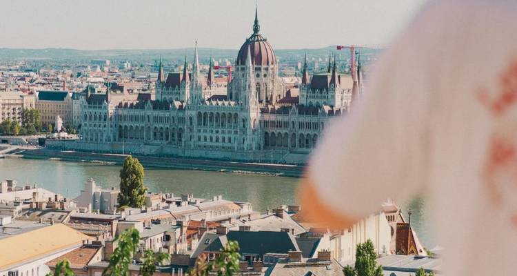 Vue panoramique diurne du Parlement de Budapest à travers le Danube, partiellement encadré par un passant au premier plan.
