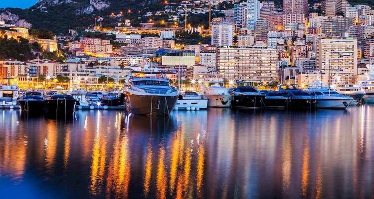 Les yachts de luxe et l'horizon vallonné de Monaco scintillent avec des lumières colorées se reflétant dans le port la nuit.