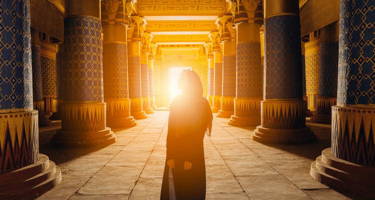 Un viajero recortado contra la luz se encuentra entre altas columnas decoradas de un salón de estilo antiguo débilmente iluminado mientras la luz solar dorada inunda a través de la puerta.