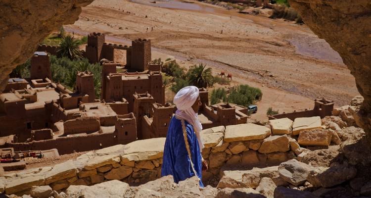 Un hombre bereber con túnica azul y turbante contempla el pueblo de barro fortificado de Ait Benhaddou desde una cueva rocosa en lo alto del valle desértico.
