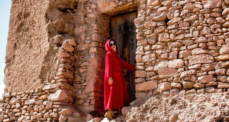 Una mujer con una capa roja con capucha está parada junto a una puerta de madera desgastada en las paredes de piedra áspera de una kasbah del desierto.