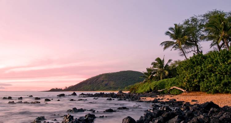 Zonsondergang op een rotsachtig strand met palmbomen en heuvels op de achtergrond.