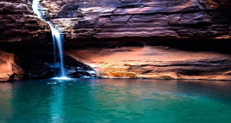 Cascada delgada cayendo en cascada hacia una piscina natural esmeralda profunda enmarcada por paredes de roca roja estratificada en Karijini.