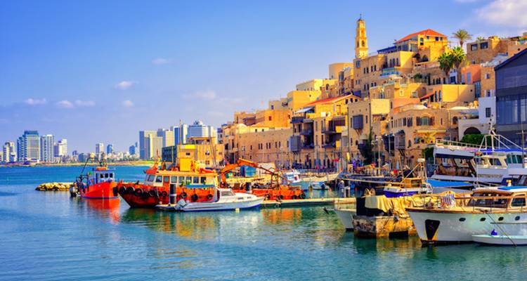 Farbenfrohe Boote im historischen Hafen von Jaffa vertaut, mit der modernen Skyline von Tel Aviv im Hintergrund.