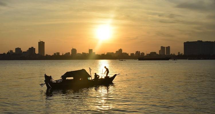 Silhouet van een persoon op een boot op een rivier tijdens zonsondergang met stadshorizon.