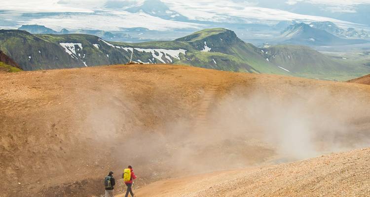 Dos excursionistas cruzan una ladera geotermal humeante con glaciares y montañas en la distancia brumosa.