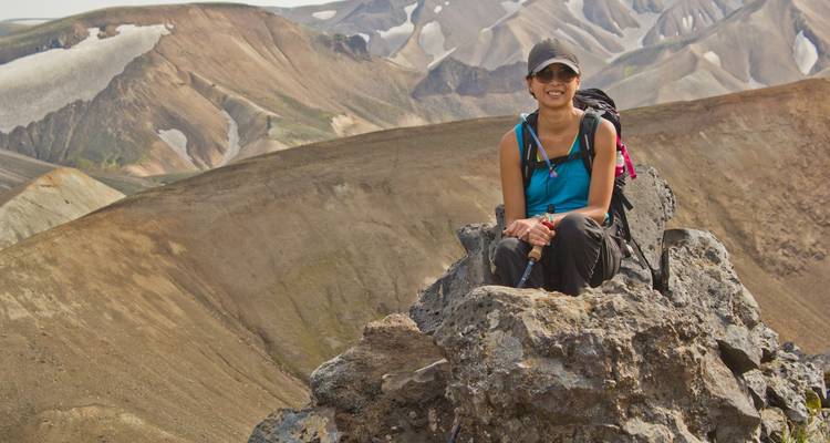 Una excursionista sonriente sentada en una formación rocosa con imponentes crestas volcánicas detrás de ella.