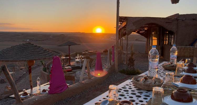 Terraza del desierto al aire libre preparada para cenar con cojines coloridos y una puesta de sol resplandeciente sobre dunas onduladas.