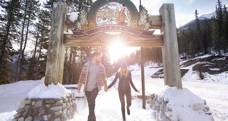 Couple walking through a snowy park with a wooden sign framing them.