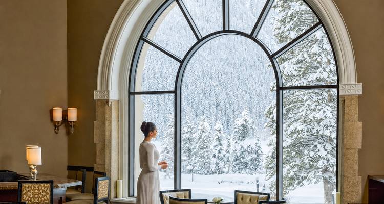 Femme dans un élégant espace de restauration regardant par une grande fenêtre en arche une forêt enneigée.