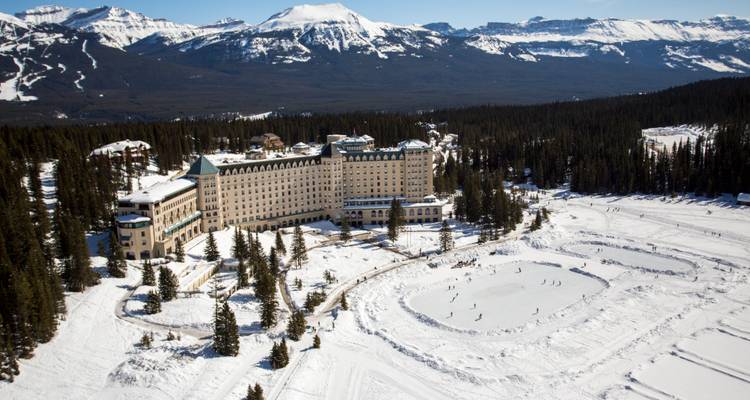 Vue aérienne hivernale du Château Lake Louise adjacent à un lac gelé entouré de patineurs et de forêt de pins noirs.