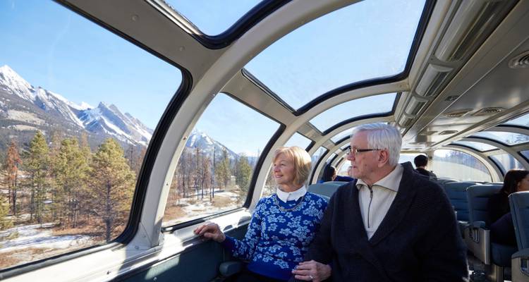 Couple de personnes âgées assis dans un train à toit de verre admirant les pics montagneux enneigés et les forêts de pins.