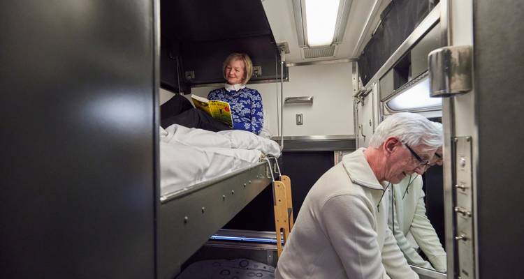 Cabine de train-couchette où une femme âgée lit sur une couchette supérieure tandis qu'un homme range ses affaires en dessous.