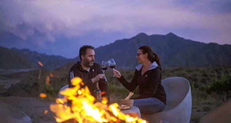 Un couple trinquant avec des verres de vin près d'un feu de camp dans un cadre montagneux.