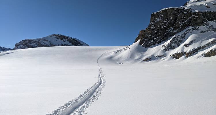Einsame Skipiste, die sich über ein großes unberührtes Schneefeld in Richtung felsiger Alpengrate erstreckt.