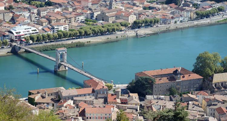 Aerial view of a suspension bridge spanning a turquoise river with red-roofed town buildings on both banks.