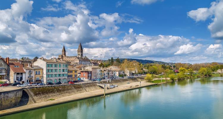 Picturesque riverside town with twin-towered Romanesque abbey reflected in calm water under patchy clouds.