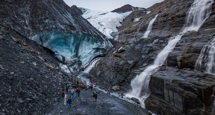Aventureros de pie en terreno rocoso cerca de una cueva de glaciar azul y cataratas en cascada.