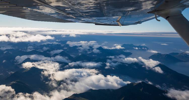 Vista aérea de picos cubiertos de nubes y el océano vistos desde el ala de un pequeño avión.