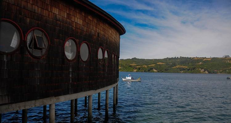 View of a circular building on stilts over water with a boat nearby.
